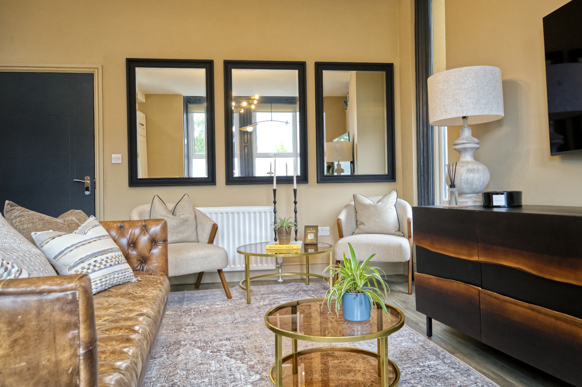 Summer suite lounge at a Soulful Stays serviced apartment in Darlington, with a leather sofa, two cream upholstered armchairs, a round coffee table, three black-framed wall mirrors and a warm lamp